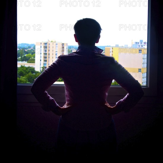 A woman stands by a window, hands on her hips, looking out at the cityscape of France. The bright daylight highlights her silhouette against the urban backdrop, creating a moment of contemplation