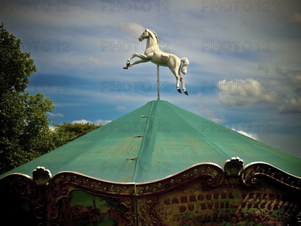 Paris. A carousel topped with a striking white horse in mid-leap stands under a vivid green canopy amidst lush trees and a bright sky, capturing the essence of joyful amusement. France
