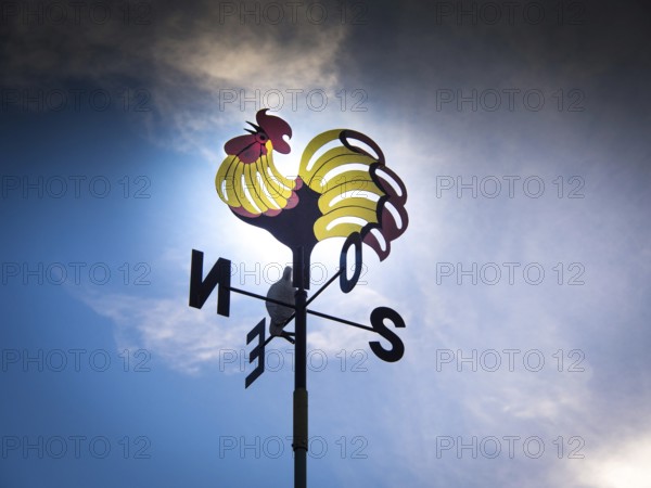 A weathercock featuring a bright rooster design sits high on a pole, silhouetted against a dramatic sky. Auvergne. France
