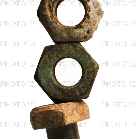 Three worn nuts and a rusty bolt are arranged vertically, highlighting their aged surfaces and unique textures. The composition emphasizes their weathered appearance against a plain backdrop