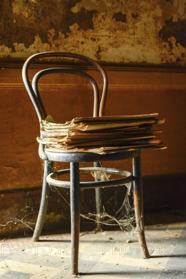 An old wooden chair is prominently positioned with a pile of yellowed newspapers resting on the seat