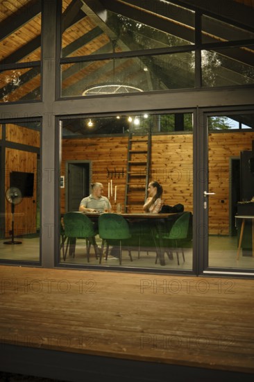 A modern wooden cabin, viewed from outside in the evening. Through large glass windows and doors a couple is seen sitting in well-lit room and talking