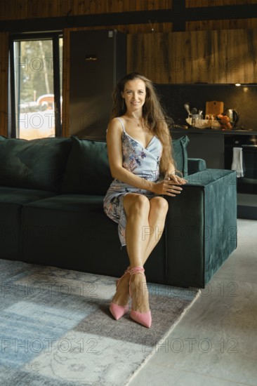 Stylish woman in floral dress seated on plush sofa in modern living room in wooden cabin