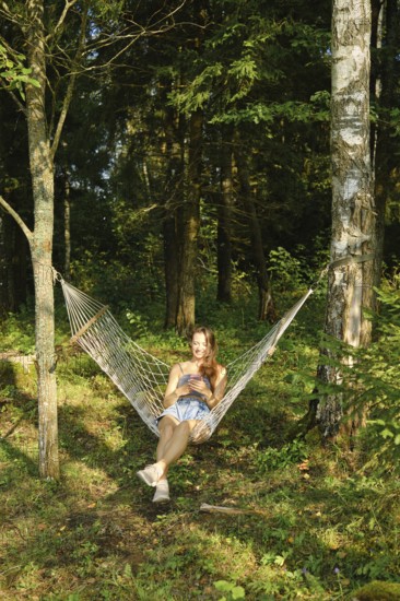 A relaxed woman reads message on smartphone while lounging in a hammock amidst lush green woods