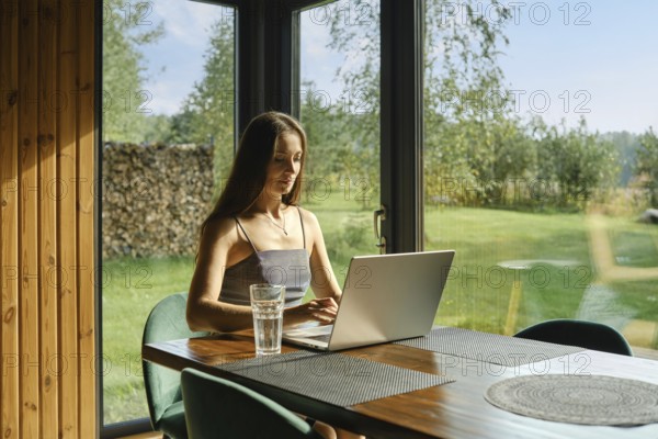 A focused woman working on a laptop in a calm cabin environment with a view of nature