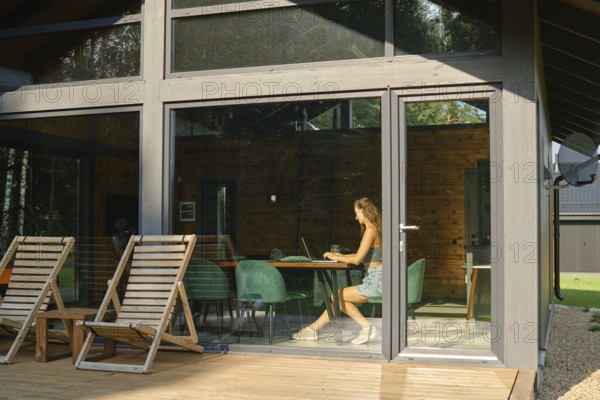 A woman is inside a modern country house with large windows and watches a TV series on a laptop while sitting at a large dining table