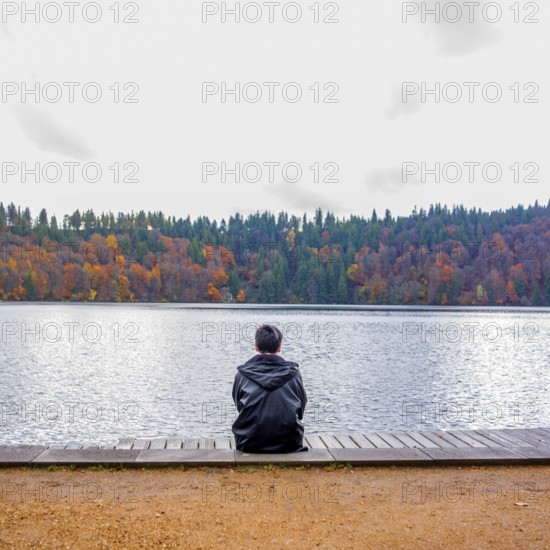 A solitary figure sits on a wooden dock, looking out over a tranquil lake. Autumn trees reflect their vibrant colors on the still water, creating a peaceful atmosphere perfect for reflection. Auvergne. France