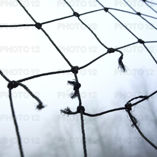 A close-up view of worn netting with frayed edges, suspended in a misty landscape. The atmosphere conveys a tranquil yet eerie feeling, suggesting neglect in a natural environment