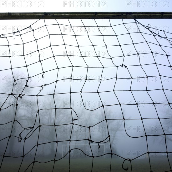 A weathered football goal net looms in the foreground surrounded by dense fog, with a blurred view of the sports field and trees in the background, creating a serene atmosphere