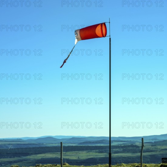 A windsock stands prominently on a pole in Auvergne, gently blowing in the breeze. The clear blue sky serves as a backdrop to rolling hills and scenic landscapes in the distance. France