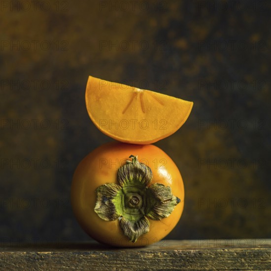 A close-up displays ripe persimmons with a vibrant orange hue, one persimmon features a slice balanced on top, highlighting its smooth skin and detailed stem, set against a dark background
