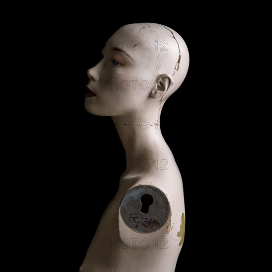 A head of a broken woman dummy is positioned in profile. The damaged surface reveals cracks and marks against a stark black background, emphasizing its worn features