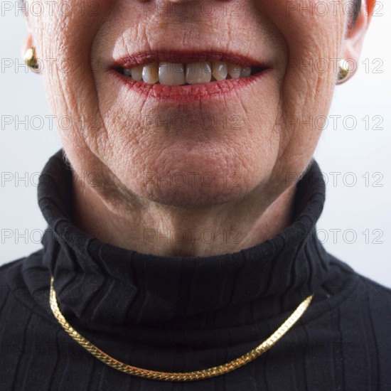 An adult woman is smiling warmly, displaying her confidence and joy. She wears a black turtleneck and a gold necklace, with her bright smile highlighted against a soft backdrop