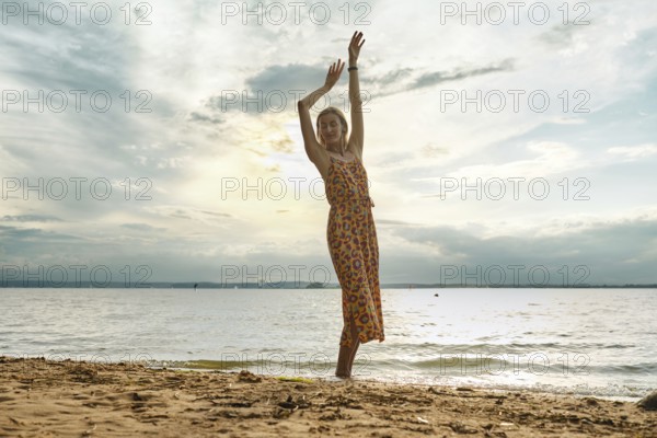 A woman in a floral sundress stands barefoot on the sand, joyfully lifting her arms. The sun sets behind her, casting a warm glow over the calm water and clouds in the sky