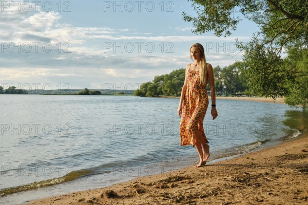 A woman in a colourful sundress strolls barefoot along the sandy shore of a tranquil lake. Lush green trees frame the water under a bright blue sky with fluffy clouds