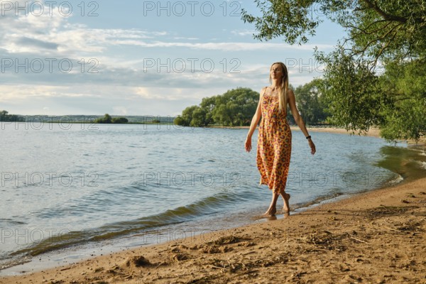 A woman in a vibrant sundress strolls barefoot along the sandy beach beside a calm lake. Sunshine illuminates the landscape, creating a peaceful atmosphere surrounded by lush greenery