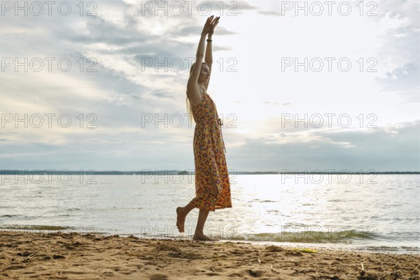 A woman in a colourful sundress walks along a serene lake, raising her arms in joy. The cloudy sky casts a soft light as waves gently lap at the sandy shore, creating a tranquil atmosphere