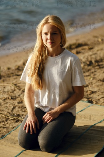 A young woman sits cross-legged on a mat at the beach, gazing thoughtfully towards the water. The calm waves reflect the soft light of the rising sun, creating a tranquil atmosphere