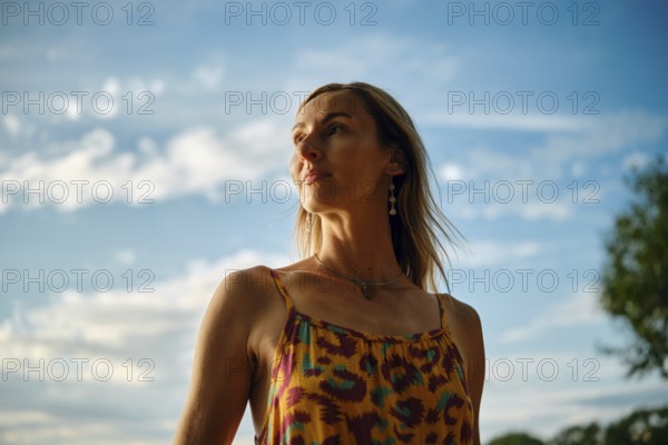 A woman stands outdoors, wearing a vibrant, colourful dress. She gazes into the distance as the sun sets, casting a warm glow on her skin. The sky is painted blue with clouds