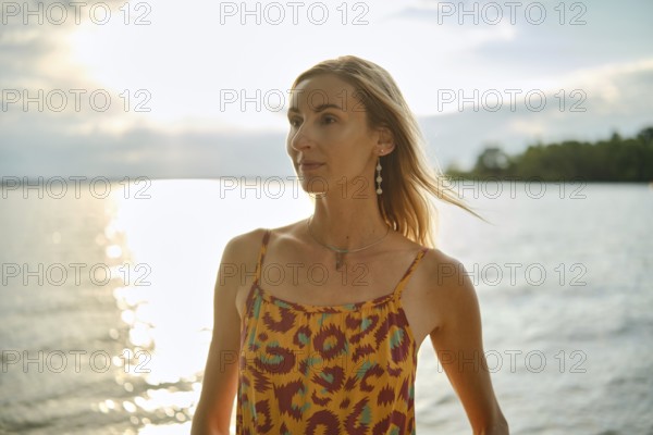 A woman with long hair poses by a calm body of water during sunset, wearing a vibrant patterned dress. Sunlight glimmers on the surface, creating a tranquil and captivating ambiance