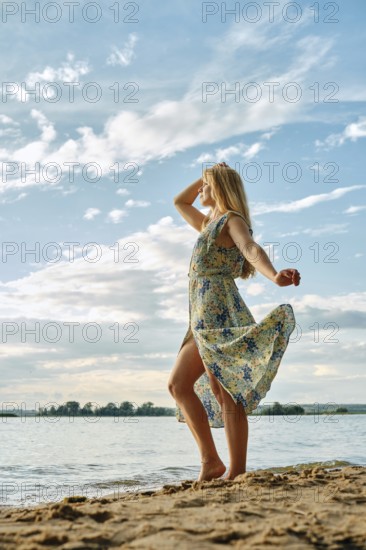 A woman stands gracefully on the sandy shore, her floral dress flowing in the wind. She gazes thoughtfully at the lake under a bright sky filled with clouds, creating a serene atmosphere