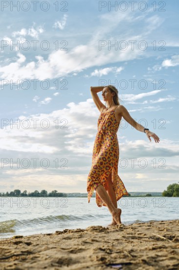 A woman stands barefoot on sandy shores, wearing a bright dress with a bold floral pattern. She playfully raises her arm against the sky with fluffy clouds during a warm afternoon