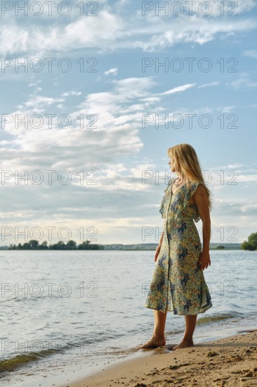A woman wearing a floral dress stands barefoot on the sandy shore of a lake, gazing at the water. The sky is partly cloudy, with soft sunlight illuminating the scene, creating a serene atmosphere