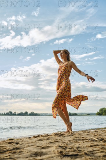 A woman twirls joyfully along the sandy shoreline, wearing a colorful dress. The sun shines brightly overhead while fluffy clouds float in the blue sky, reflecting a leisurely, carefree summer day