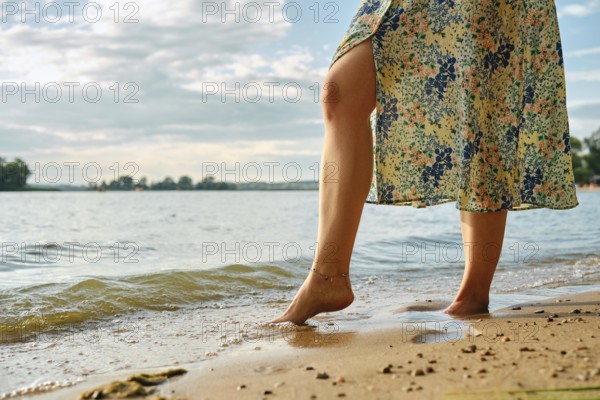 Unrecognizable woman stands at the edge of a lake, her bare foot touching the gentle waves at sunset. She wears a flowery dress, and the sun casts a warm glow on the serene landscape