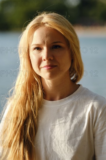 A woman with long flowing hair stands by a serene body of water. Her relaxed expression showcases joy in the sunlight as she enjoys a peaceful afternoon outdoors surrounded by nature
