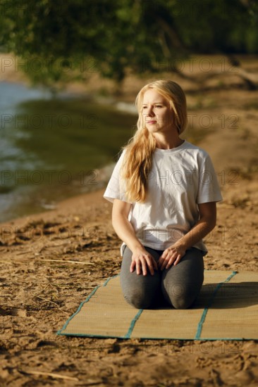 A woman sits on a mat by the riverside, engaged in a moment of mindfulness. She has long blonde hair and wears a simple white t-shirt and gray leggings. The warm sun illuminates the peaceful setting