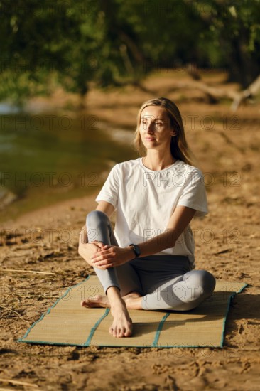A woman sits cross-legged on a mat by a riverbank, focusing on mindfulness as the sun sets. She enjoys the serene environment, surrounded by nature and reflecting on her thoughts