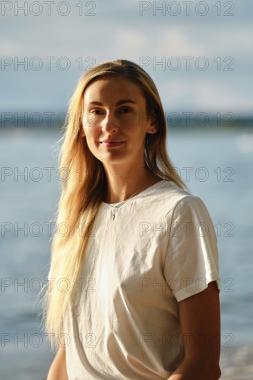 A woman with long hair smiles gently while standing near the water's edge at the beach. The bright sunlight highlights her face, and the waves create a serene backdrop on a warm day