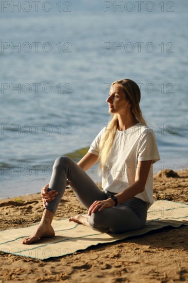 A woman sits on yoga mat by the lakeshore and listens a surf noise. The calm water reflects the evening light as she sits gracefully, enjoying the peaceful ambiance and gentle breeze
