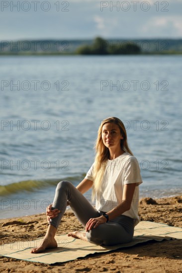 A woman sits peacefully on a mat by the lake, enjoying the serene atmosphere as the sun sets. She is focused on her thoughts while surrounded by tranquil waters and natural beauty