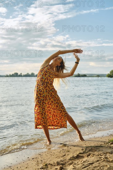 A woman dressed in a vibrant, patterned dress dances gracefully near the edge of a lake. The sun shines brightly, casting a warm glow over the calm waters and sandy shore, creating a serene ambiance
