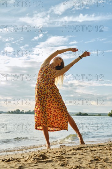 A joyful woman stretches gracefully on the sandy shore as the sun sets over the water. The vibrant dress she wears flows with her movements, capturing the serene beauty of the beach