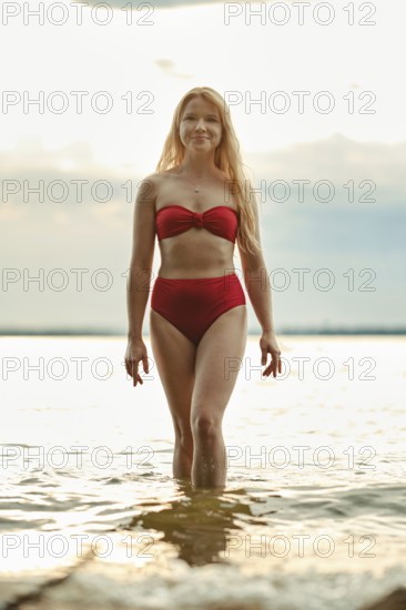 A woman with long hair walks confidently in shallow water, wearing a red bikini. The sun sets in the background, casting a warm glow over the beach. It is a serene summer evening by the lake
