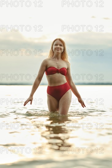 A young woman with long hair walks through shallow water, showcasing a vibrant red swimsuit. The soft light from the evening sky creates a serene atmosphere at a lakeside setting