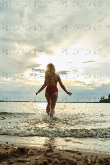A woman in a red swimsuit steps into a tranquil lake, creating splashes as the sun sets behind her. The sky is filled with soft clouds reflecting warm colors, enhancing the serene atmosphere