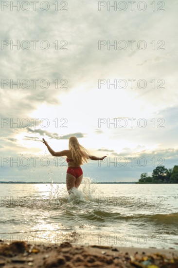 A woman in a red swimsuit plays in the shallow water of a lake. The sun sets behind her, casting a warm glow while she joyfully splashes and embraces the tranquil moment