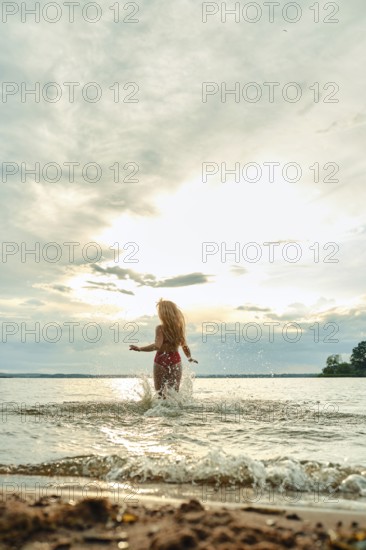 A woman with long hair playfully splashes in the water at a beach during sunset. The sky is partially cloudy, and the sun is setting, creating a warm glow over the scene. The water is calm
