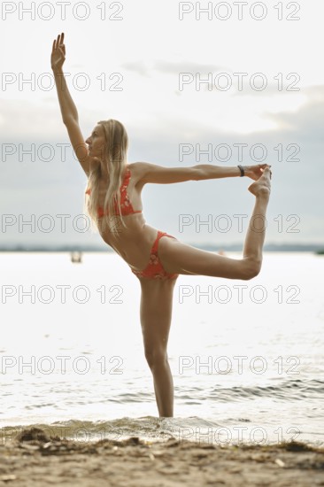 A dancer showcases her flexibility and grace by striking an impressive pose at the water's edge. The sunset casts a soft glow, enhancing the tranquil atmosphere of the lakeside setting
