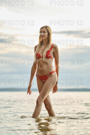 A woman strolls gracefully through shallow waters at sunset, wearing a vibrant red bikini adorned with floral patterns. The tranquil lake reflects the cloudy sky and creates a serene atmosphere