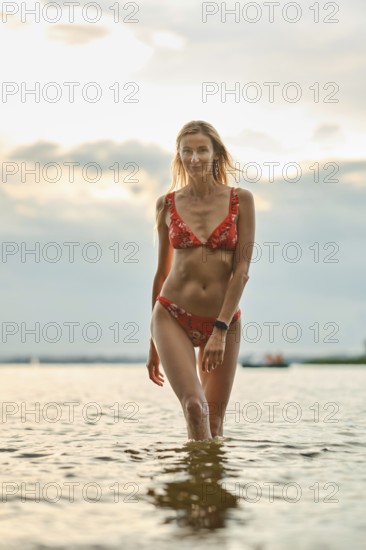 A woman strolls through shallow water as the sun sets behind her. Dressed in a floral bikini, she enjoys the warm evening while reflecting on the peaceful surroundings of the lake