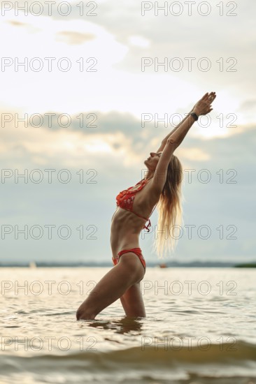 A woman in a red bikini stands in profile knee-deep in a calm lake with arms raised high, embracing the sunset. Her long hair flows as she enjoys the serene atmosphere, surrounded by soft clouds