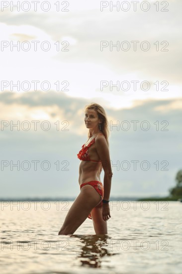 A woman in a red bikini walks through calm waters, silhouetted against a sunset with clouds overhead. She enjoys the serene atmosphere of the lakeshore, creating a peaceful moment of relaxation