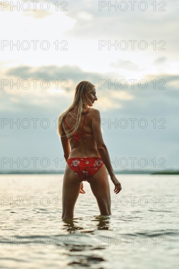 A woman stands in waist-deep water, facing away, as the sun sets behind her. She wears a vibrant red bikini with floral patterns, accentuating the natural beauty of the lakeside setting