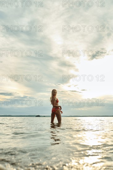 A woman stands in shallow water, enjoying the tranquil surroundings as the sun sets on the horizon. She looks back and calls out to join her