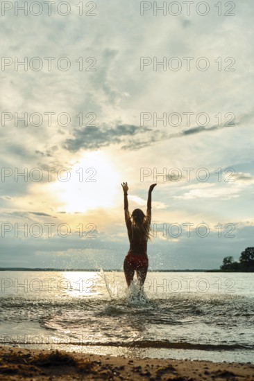 A woman stands in shallow water at a beach during sunset, joyfully raising her arms towards the colorful sky. The sun casts warm hues on the horizon, reflecting on the water's surface